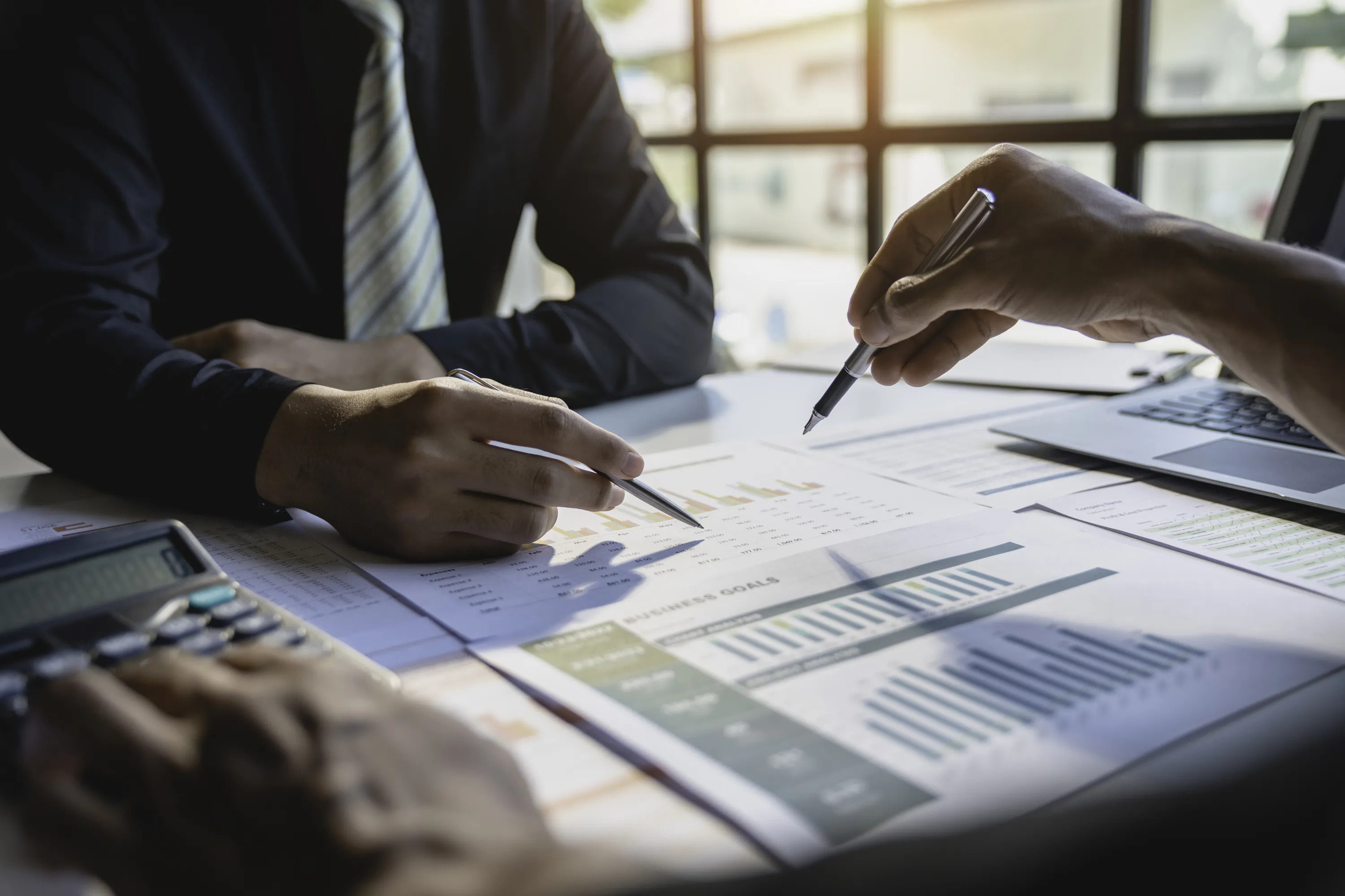 Kaksi henkilöä tarkastelee liiketoimintatavoitteita ja kaavioita pöydällä, jossa on laskin ja kannettava tietokone, valaistus on kirkas.